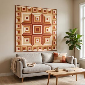 May include: A large, geometric quilt in shades of brown, tan, and cream hangs on a wall above a gray sofa. The quilt features a square pattern with concentric squares and rectangles. A matching pillow and a wooden coffee table complete the living room scene.