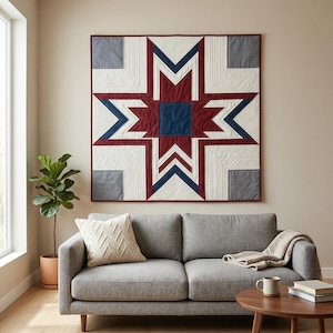 May include: A large, geometric quilt with a starburst design in shades of red, white, blue, and gray hangs on a wall. The quilt is above a gray sofa with a cream-colored pillow. A small, round wooden table and a potted plant are in the foreground.