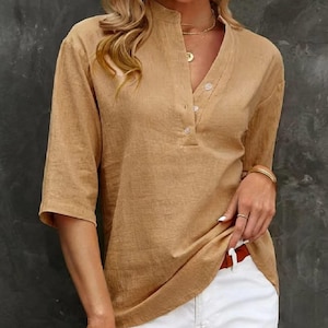 May include: A tan linen shirt with a mandarin collar and a V-neck. The shirt features three buttons and three-quarter sleeves. The model is wearing the shirt with white trousers and gold jewellery.