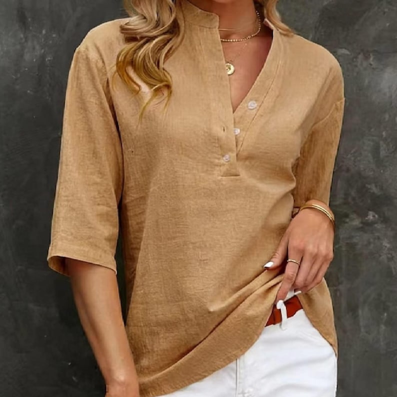 May include: A tan linen shirt with a mandarin collar and a V-neck. The shirt features three buttons and three-quarter sleeves. The model is wearing the shirt with white trousers and gold jewellery.