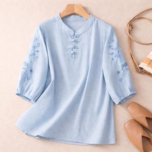 May include: A pale blue blouse with three-quarter length sleeves and a mandarin collar. The blouse has blue floral embroidery on the sleeves and fabric-covered buttons down the front. The top is displayed on a wooden hanger.