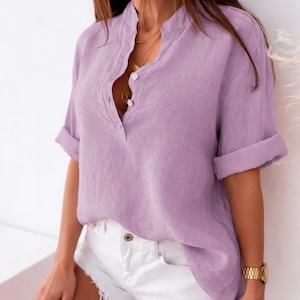 May include: A lilac linen shirt with a mandarin collar and rolled sleeves. The shirt features a button-down front and is worn with white shorts. A gold watch is visible on the wrist.