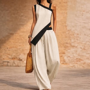 May include: A two-piece outfit consisting of a sleeveless, off-white top with a diagonal black panel and matching wide-leg trousers. The top features a high neckline and a side cutout. A woven tan handbag is held at the side of the garment.
