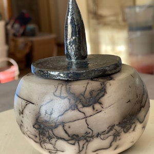Op de afbeelding: Een handgemaakte keramische pot met deksel. De pot is rond met een crèmekleurige basis en ingewikkelde zwarte craquelé patronen. Het deksel en de conische handgreep zijn donkergrijs. Een uniek decoratief stuk.