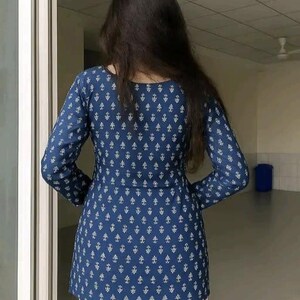 May include: A long-sleeved, navy blue tunic with a white geometric pattern. The tunic is paired with white pants. The back view of the outfit is shown, with the person facing away from the camera.