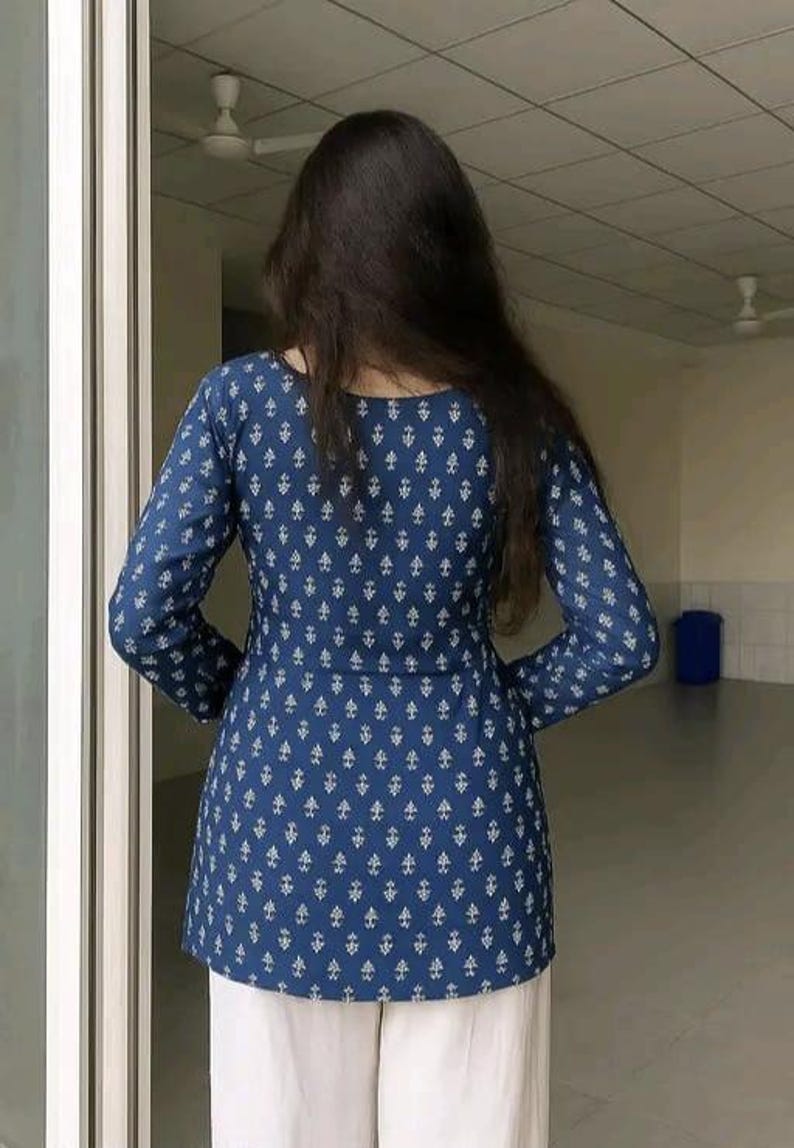 May include: A long-sleeved, navy blue tunic with a white geometric pattern. The tunic is paired with white pants. The back view of the outfit is shown, with the person facing away from the camera.