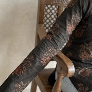 May include: Dark charcoal-colored long-sleeved top with a floral pattern in a reddish-brown hue. The top is worn by a person sitting on a wooden chair. The floral design is repeated on the sleeves and body of the top.