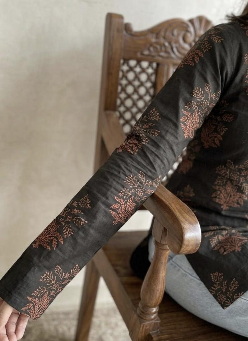 May include: Dark charcoal-colored long-sleeved top with a floral pattern in a reddish-brown hue. The top is worn by a person sitting on a wooden chair. The floral design is repeated on the sleeves and body of the top.