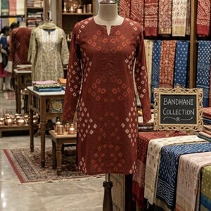 Puede incluir: Una t&uacute;nica granate de manga larga con un estampado de diamantes sobre un maniqu&iacute;. La t&uacute;nica se exhibe en una tienda con otras prendas y textiles. Un letrero dice "BANDHANI COLLECTION".