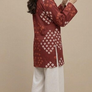 Puede incluir: Una mujer con una t&uacute;nica granate con un estampado geom&eacute;trico blanco, combinada con pantalones blancos de pernera ancha. Sostiene un smartphone. Un peque&ntilde;o bolso de mano tejido descansa a sus pies. El atuendo es adecuado para ropa informal.