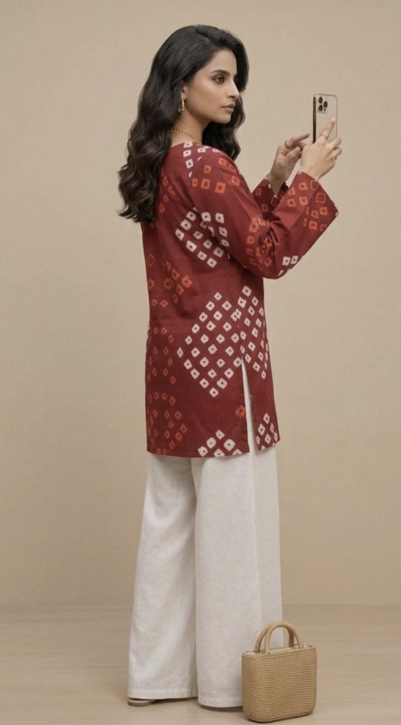 Puede incluir: Una mujer con una t&uacute;nica granate con un estampado geom&eacute;trico blanco, combinada con pantalones blancos de pernera ancha. Sostiene un smartphone. Un peque&ntilde;o bolso de mano tejido descansa a sus pies. El atuendo es adecuado para ropa informal.