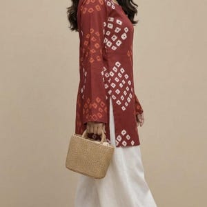 Puede incluir: Conjunto femenino que incluye una t&uacute;nica burdeos de manga larga con un estampado geom&eacute;trico blanco, combinada con pantalones blancos de pernera ancha. Un peque&ntilde;o bolso de mano tejido completa el conjunto. Adecuado para ocasiones informales o semiformales.