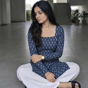 May include: A woman wearing a blue floral print long-sleeve top and white pants. The top has a square neckline and the pants are wide-legged. She is wearing a gold watch and black sandals. The outfit is casual and comfortable.