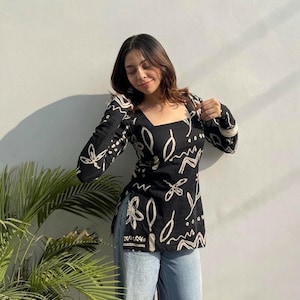 Pode incluir: Top preto de manga comprida com decote quadrado e padrões abstratos brancos de flores e linhas. O top é combinado com jeans azul claro. A roupa é usada em frente a uma parede branca com uma planta verde no canto.