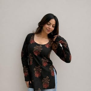 May include: Black long-sleeve tunic with a scoop neck and a floral pattern in shades of red and gold. The tunic is paired with light blue jeans. The floral design is repeated on the sleeves.