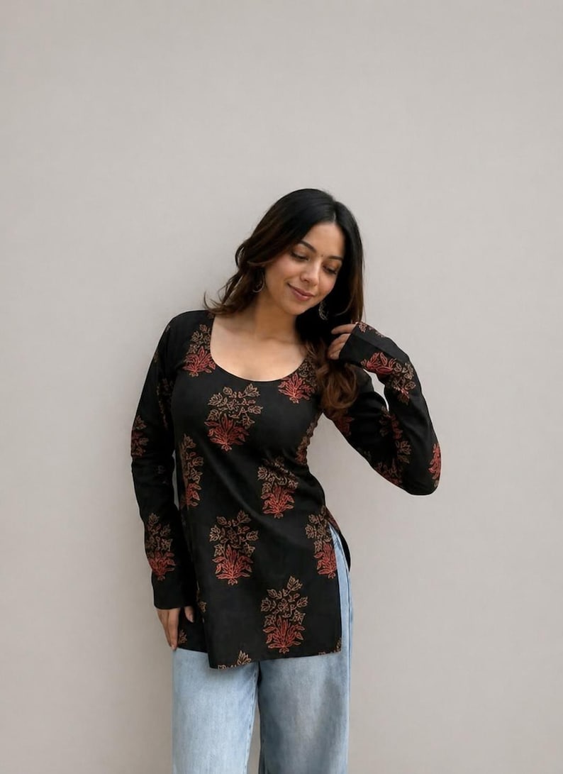May include: Black long-sleeve tunic with a scoop neck and a floral pattern in shades of red and gold. The tunic is paired with light blue jeans. The floral design is repeated on the sleeves.