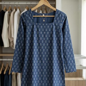 May include: A blue tunic with a square neckline and long sleeves, adorned with a white floral pattern. The tunic is hanging on a wooden hanger, with a gold necklace. The background shows a closet with clothes.