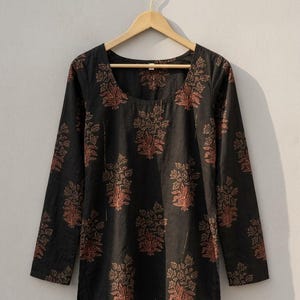 May include: Black long-sleeve tunic with a scoop neckline, adorned with a repeating floral pattern in shades of rust and gold. The tunic is hanging on a wooden hanger against a neutral background. This garment is suitable for casual wear.