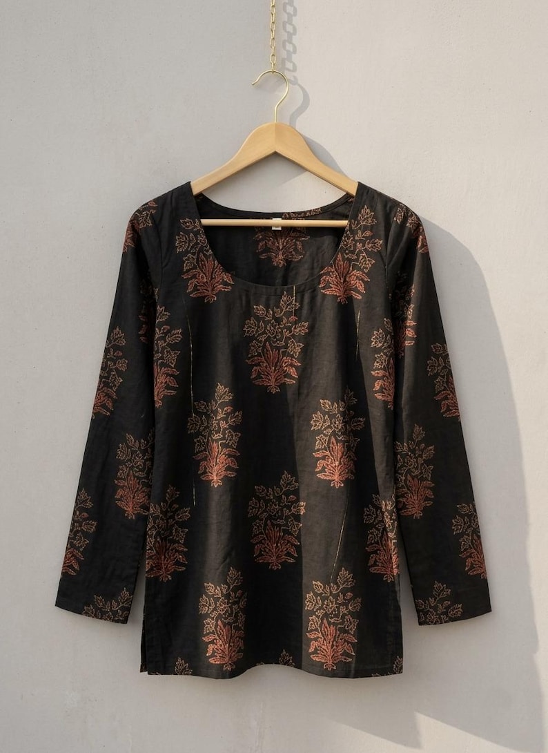 May include: Black long-sleeve tunic with a scoop neckline, adorned with a repeating floral pattern in shades of rust and gold. The tunic is hanging on a wooden hanger against a neutral background. This garment is suitable for casual wear.