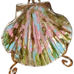 Könnte beinhalten: Ein dekoratives Jakobsmuschel-Objekt mit handbemalter Oberfläche, die Streifen in Pastellrosa, Hellblau und Metallic-Gold aufweist. Die Muschel wird auf einem goldfarbenen Metalldrahtständer mit geschwungenen Füßen präsentiert.