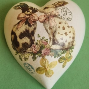 Puede incluir: Adorno decorativo en forma de corazón con dos conejos con lazos rosas, rodeados de flores. Un conejo es blanco y negro, el otro blanco con manchas marrones. Estilo vintage con sellos y texto.