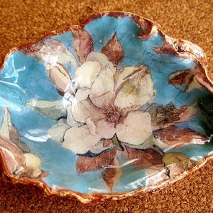 May include: A decorative, handmade, blue and gold-edged dish with a floral design. The dish features a large, white and pink flower in the center, surrounded by brown and green leaves. The dish is approximately 6 inches in diameter.