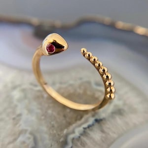 May include: A gold ring shaped like a snake, with a red gemstone eye. The snake's body is detailed with small, round beads. The ring is a piece of jewellery, likely worn on a finger.