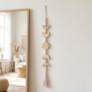 Op de afbeelding: Een verticale wandhanger met een reeks lichtgekleurde houten maanfasen, waaronder halve manen en een volle cirkel, gescheiden door kleine houten kralen aan een koord. Het object eindigt onderaan met een lichtgekleurde stoffen kwast.