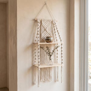 Può includere: Una mensola a muro in macramè con due ripiani in legno. La mensola è sospesa da un design in macramè intrecciato ed è appesa a una parete beige. Un piccolo vaso di ceramica e un vaso di vetro con verde sono sui ripiani.