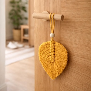May include: A mustard yellow macrame leaf hanging from a wooden door handle. The leaf has a detailed woven pattern and a small wooden bead. The door and handle are made of light-colored wood.