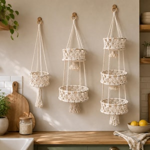 Peut inclure: Trois paniers suspendus en macramé blanc cassé accrochés à des anneaux en bois. Le modèle de gauche a un étage, celui du centre deux étages et celui de droite trois étages. Chaque panier présente des nœuds complexes, des cercles en bois et des pompons.