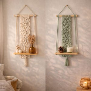 May include: Two macrame hanging shelves, one cream and one sage green, each with a wooden shelf. The cream shelf holds a brown vase with dried flowers, while the green shelf displays a pinecone and a candle. Both shelves have a leaf pattern.