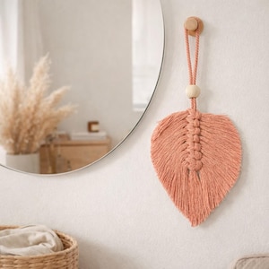 Op de afbeelding: Perzikroze macramé blad wandhanger. Het bladontwerp is opgehangen aan een houten peg met een geknoopt koord en een houten kraal. Het blad heeft een getextureerde, gefranjerde uitstraling en hangt aan een witte muur.