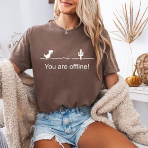 May include: A brown t-shirt featuring a white graphic of a dinosaur, cactus, and the text "You are offline!". This casual t-shirt is suitable for everyday wear.