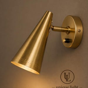 May include: A brass wall-mounted lamp with a conical shade and a round base. The lamp has a small switch and a pivoting arm. The shade has a series of small holes near the bottom. The text "unique light" is below a logo.