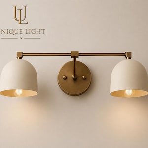May include: A two-light wall sconce with a brass finish and off-white, dome-shaped shades. The sconce features a circular backplate and a horizontal bar connecting the two lights. The text "UNIQUE LIGHT" is at the top.