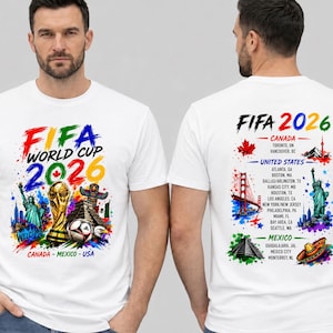 May include: A white t-shirt featuring a FIFA World Cup 2026 graphic. The front displays the tournament logo, a soccer ball, the Statue of Liberty, and a pyramid. The back lists host cities in Canada, the United States, and Mexico with icons.