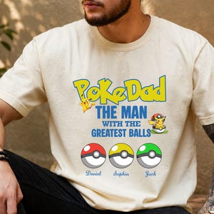 May include: Cream-colored t-shirt featuring the text "PokeDad" in yellow and blue, with Pikachu graphics. Below, it reads "THE MAN WITH THE GREATEST BALLS." Three Poke Balls are below with names: Daniel, Sophia, and Jack. The shirt is a casual wear item.