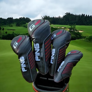 Puede incluir: Un juego de fundas negras para palos de golf con la marca "PING" en letras blancas. Las fundas tienen líneas rojas y el logotipo "G410". El fondo muestra un campo de golf con hierba verde y árboles.