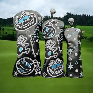 May include: Set of three golf club head covers in black with whimsical cartoon designs. Each cover features a different design, including a grinning cat wearing sunglasses, a bomb with a smiley face, and stars. The covers are set against a green golf course.