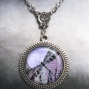 May include: Silver-toned necklace with a round pendant featuring a dragonfly design. The pendant has a lavender moon and a dragonfly silhouette over a background of script and tall grass. The necklace has a decorative bail and chain.