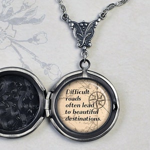 May include: Silver locket necklace with a compass and the quote "Difficult roads often lead to beautiful destinations." The locket is open, revealing a black patterned interior.