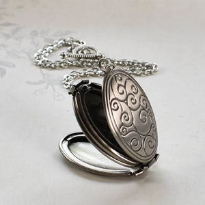 May include: A silver oval locket necklace with a swirling pattern on the front. The locket is open, revealing a black interior.