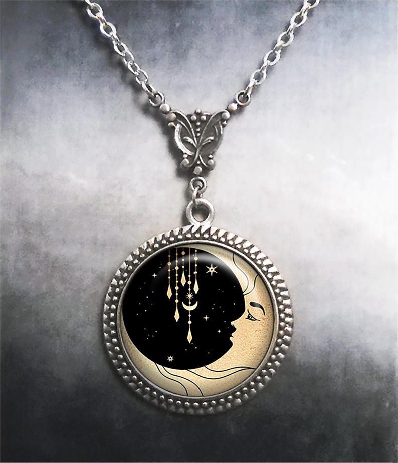 May include: A silver necklace with a round pendant featuring a crescent moon with a face and a starry night sky. The pendant has a gold-colored design of a crescent moon and stars hanging from the top.