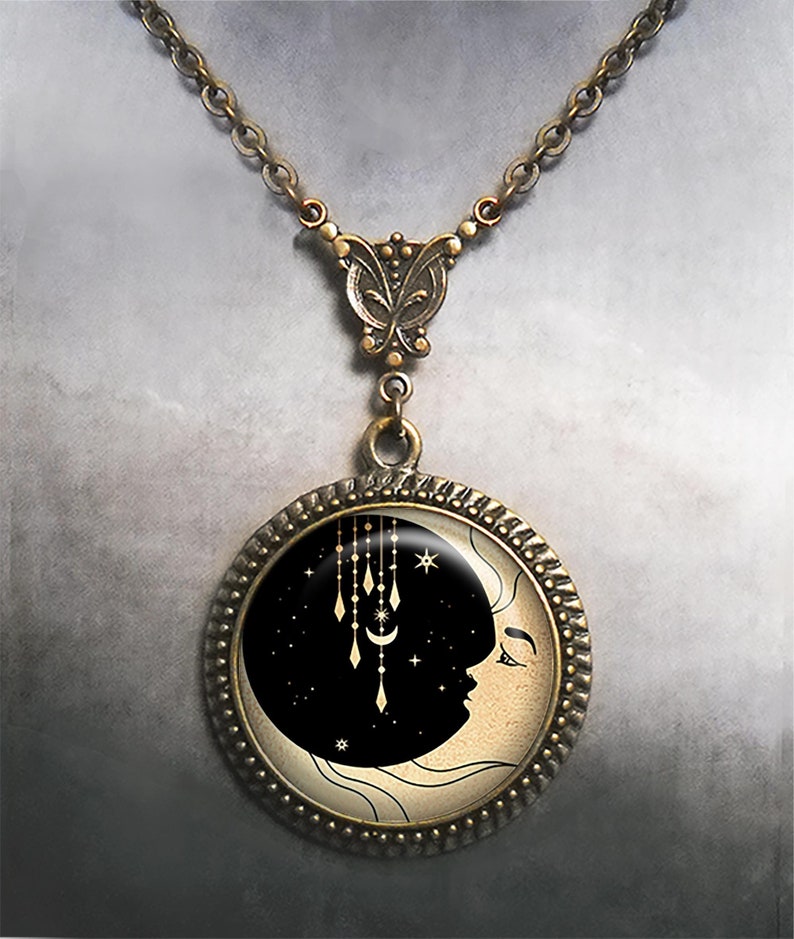 May include: A gold-toned necklace with a round pendant featuring a black crescent moon with a face and gold stars and dangling charms against a cream background.