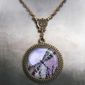 May include: A bronze-toned necklace featuring a round pendant with a dragonfly design. The pendant's image includes a full moon, script, and tall grass. The necklace has a butterfly-shaped clasp and a chain.