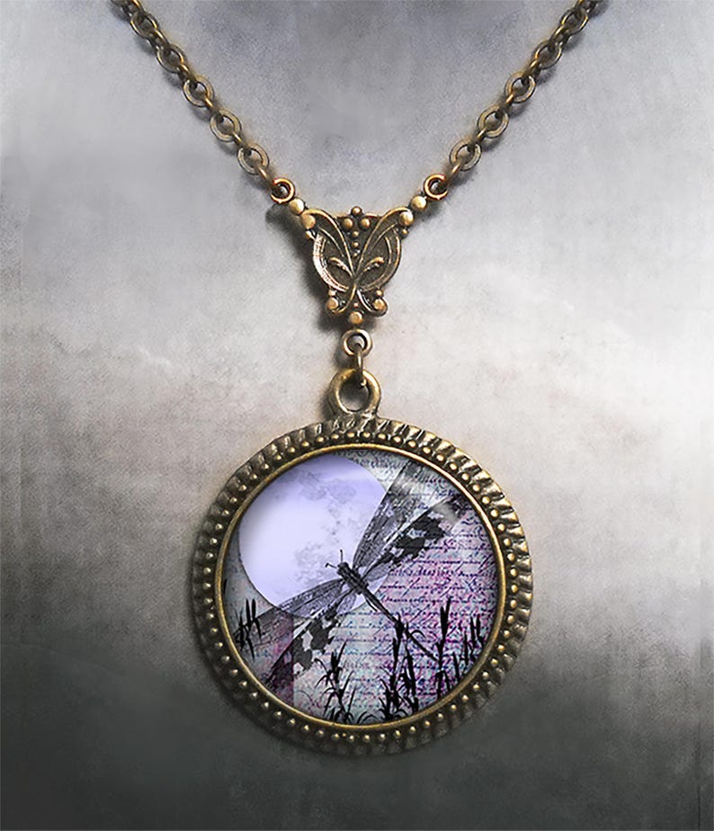 May include: A bronze-toned necklace featuring a round pendant with a dragonfly design. The pendant's image includes a full moon, script, and tall grass. The necklace has a butterfly-shaped clasp and a chain.