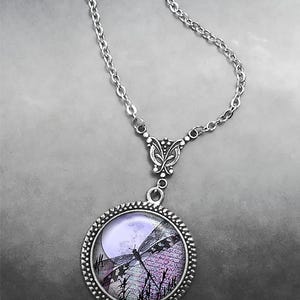 May include: Silver-toned necklace featuring a round pendant with a dragonfly design. The pendant showcases a dragonfly silhouette against a moonlit scene with purple and black hues. The chain is a delicate link style.