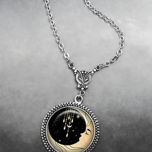 May include: A silver chain necklace with a round pendant featuring a black crescent moon with a face and gold stars and dreamcatchers. The pendant is surrounded by a silver beaded border.