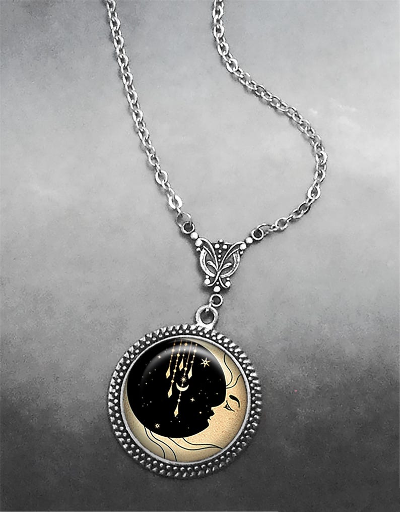 May include: A silver chain necklace with a round pendant featuring a black crescent moon with a face and gold stars and dreamcatchers. The pendant is surrounded by a silver beaded border.